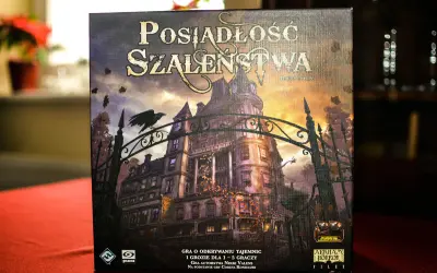 Pudełko gry "Posiadłość Szaleństwa" z mroczną posiadłością i wroną na bramie. Dodatki do tej gry planszowej zapowiadają nowe, przerażające przygody.