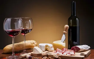Deux verres de vin rouge, une bouteille, du pain, des fromages, de la charcuterie et des noix. Un accord parfait pour un vin pour fromage et charcuterie.