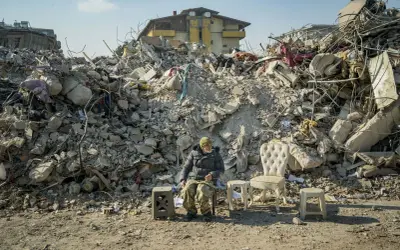 Mężczyzna siedzi wśród gruzów po trzęsieniu ziemi w Turcji. Obok niego stoją meble, jakby czekał na powrót do normalności.