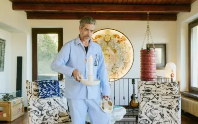 Uomo in pigiama azzurro tiene candela accesa e tazza di caffè, in un ambiente eclettico con opere d'arte e mobili bizzarri. Un'immagine che evoca un momento di riflessione, quasi un rituale mattutino, dove il lavoro incontra il sogno.