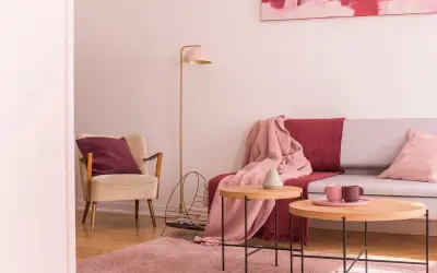 Przytulny salon z fotelem w kolorze ecru i amarantową poduszką. Na kanapie narzuta w tym samym amarantowym kolorze.