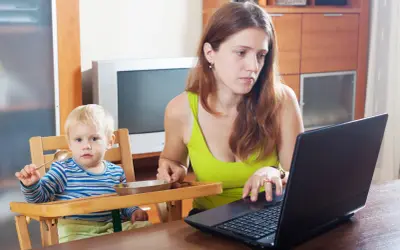 Matka z dzieckiem przy stole, zastanawia się, ile trwa urlop macierzyński, pracując na laptopie.