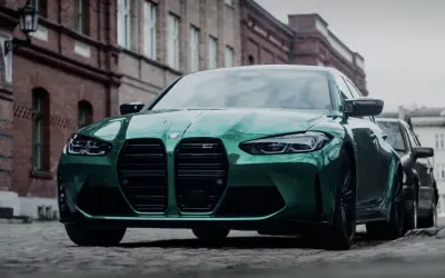 Zielony BMW M3 Competition z charakterystycznym grillem, zaparkowany na brukowanej ulicy przed ceglanymi budynkami.