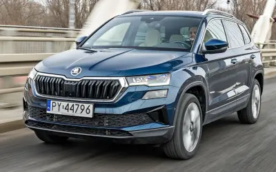Ciemnoniebieska Skoda Karoq jedzie po moście. Kierowca uśmiecha się, a za nim widać drzewa i budynki.