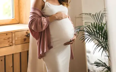 Frau in weißem Kleid und rosafarbener Strickjacke hält ihren Bauch, 17 ssw.