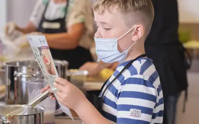Junge mit Maske liest Rezept, während andere kinder kochen selbst. Küchenutensilien und Töpfe sind zu sehen.