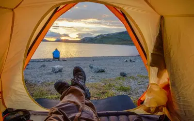 Blick aus dem Zelt auf einen See bei Sonnenuntergang. Eine Person sitzt am Ufer. Ja, darf man in Schweden wildcampen!