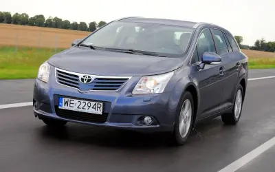 Szary kombi Toyota Avensis T27 1.8 benzyna jedzie po drodze, za nim pola i drzewa.