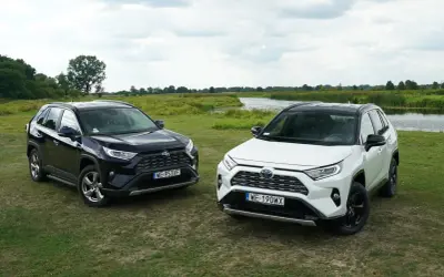 Dwa nowe modele Toyota RAV4 2025, czarny i biały, zaparkowane na trawie nad jeziorem.
