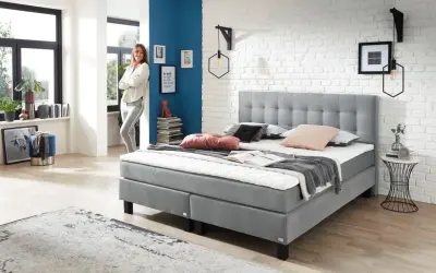 Modernes Boxspringbett in Grau, ideal für erholsamen Schlaf bei Bandscheibenvorfall. Eine Frau lehnt lässig daneben.