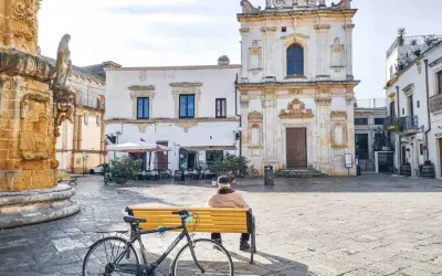 Un uomo seduto su una panchina nel **pescoluse centro storico**, con una bicicletta appoggiata. Sullo sfondo, una chiesa barocca e un caffè all'aperto.