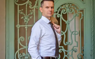 Tomasz Roy Szabelewski w białej koszuli i krawacie, z marynarką na ramieniu, przed ozdobnymi drzwiami.