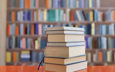Stos książek na biurku, tło biblioteki z kolorowymi grzbietami. Idealne lektury na e8.