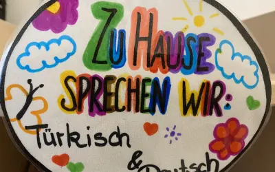 Bunte Zeichnung mit der Aufschrift "Zu Hause sprechen wir: Türkisch & Deutsch". Ein Beispiel für vorurteilsbewusste Pädagogik, die Mehrsprachigkeit feiert.