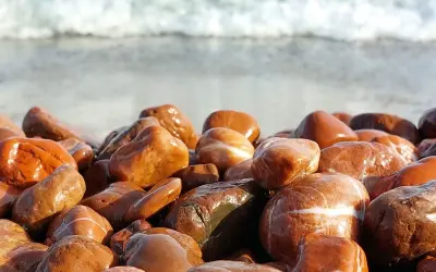 Gładkie, mokre kamienie na plaży, przypominające jaspis czerwony właściwości. W tle widać falę.
