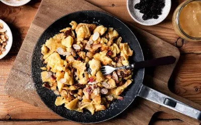 Kaiserschmarrn-Rezepte: Ein goldbrauner Kaiserschmarrn mit Mandelsplittern, Rosinen und Apfelstücken in einer Pfanne, bestäubt mit Puderzucker.