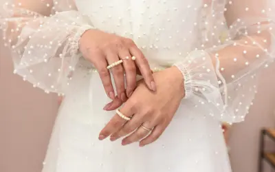 Dłonie panny młodej ozdobione pierścionkami z perłami, na tle sukni ślubnej z delikatnymi koralikami. Perły na ślubie dodają elegancji.