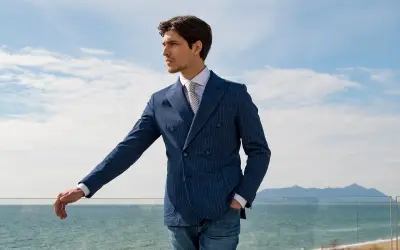 Uomo con giacca blu a doppio petto a righe, camicia bianca e cravatta, appoggiato a una ringhiera con vista mare.