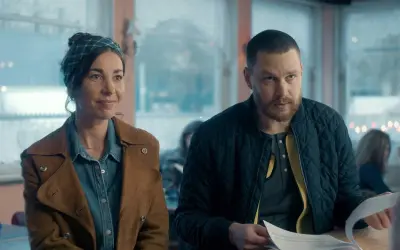 En kvinna med scarf och en man med skägg sitter vid ett bord. Scenen är från en dansk serie på SVT.