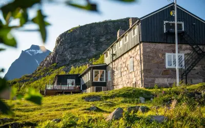 STF Kebnekaise fjällstation badar i solljus. En stenbyggnad med mörka trädetaljer reser sig mot en dramatisk bergsklippa.