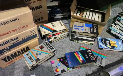 Kolekcja kaset VHS, w tym marki Movix, Konica, Maxell, Ariex, Yamaha. W tle pudełka z kasetami, przypominające o opłacie reprograficznej.