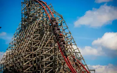 Pasażerowie Hyperion roller coaster zjeżdżają po drewnianej konstrukcji, która wznosi się ku błękitnemu niebu z chmurami.