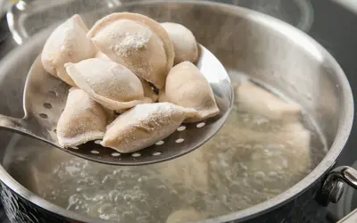 Gotowanie pierogów: na łyżce cedzakowej świeżo uformowane pierogi czekają na wrzucenie do wrzątku. W garnku bulgocze woda, gotowe do przyjęcia pysznego farszu.