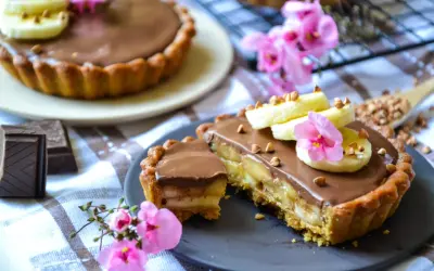 Tarte banane chocolat, garnie de rondelles de banane, de fleurs roses et de noisettes concassées. Un délice gourmand !