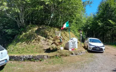 Monumento commemorativo con bandiera italiana e corona d'alloro al passo delle radici, circondato da alberi e con un'auto parcheggiata.