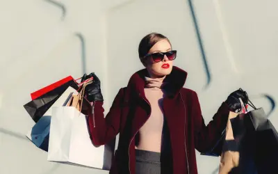 Donna con occhiali da sole e rossetto rosso, avvolta in un cappotto bordeaux, porta borse della spesa, simbolo di moda veloce.