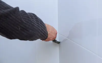 Dłoń w swetrze usuwa starą fugę silikonową z narożnika białych płytek za pomocą narzędzia.
