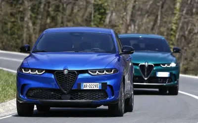 Dwa Alfa Romeo Tonale, jeden niebieski z przodu, drugi zielony z tyłu, jadą drogą. Sprawdź alfa romeo tonale cena.