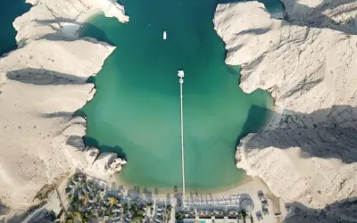 Vista aerea di un resort in Oman con bambini che giocano sulla spiaggia. Acque turchesi, montagne rocciose e un lungo pontile.