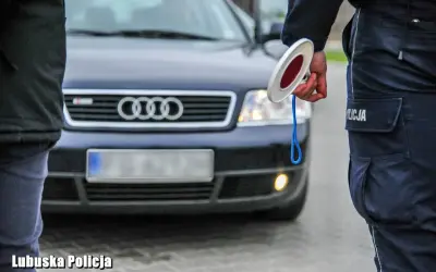 Policjant z lizakiem zatrzymuje auto. Widać jego mundur i fragment ciemnego Audi. Jakie światła podczas deszczu, gdy policja kontroluje ruch.