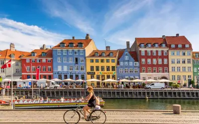 En kvinna cyklar längs Nyhavn i Köpenhamn, ett soligt exempel på danmark klimat. Färgglada hus och kanalbåtar skapar en livlig atmosfär.