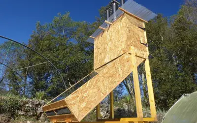 Un sechoir solaire artisanal en bois, avec un panneau incliné vitré, sous un ciel bleu.