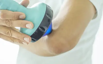 Une main utilise un appareil bleu clair pour soigner une tendinite au coude. La lumière bleue émise par l'appareil cible la zone douloureuse.