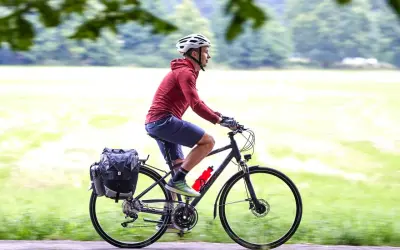 Mężczyzna w kasku jedzie na rowerze trekkingowym z sakwami, ciesząc się przejażdżką pośród zieleni.