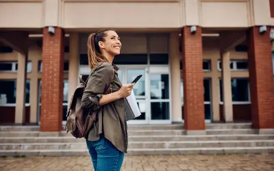 Uśmiechnięta studentka z plecakiem i telefonem przed budynkiem uczelni. Zastanawia się, jakie studia po rozszerzonym angielskim wybrać.