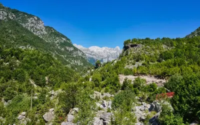Grüne Hänge und felsige Gipfel in den albanischen Alpen unter strahlend blauem Himmel. Ein kleiner roter Steg führt über einen steinigen Bachlauf.