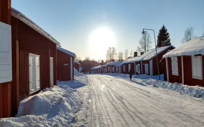 Vinterdag i kyrkbyn Gammelstad. Röda trähus med snö på taken kantar en snötäckt väg. Solen skiner starkt på en klarblå himmel.