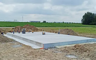 Płyta fundamentowa z wystającymi rurami na placu budowy, w tle pola i budynki.