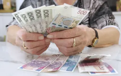 Dłonie z obrączkami trzymają banknoty 100 i 500 zł. Na stole leżą banknoty 20 zł. Czerwcowe emerytury już są!