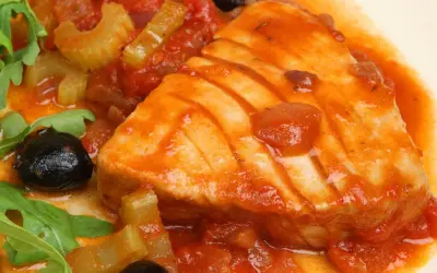 Un succulento tonno in padella con pomodoro, olive nere e sedano, guarnito con rucola fresca.