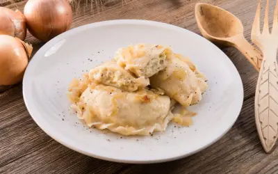 Pierogi z mięsem i cebulką na białym talerzu, obok cebule i drewniane sztućce.