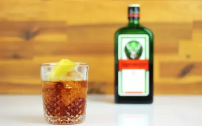 Orzeźwiający jager z colą, z plasterkiem cytryny, obok zielonej butelki Jägermeistera.