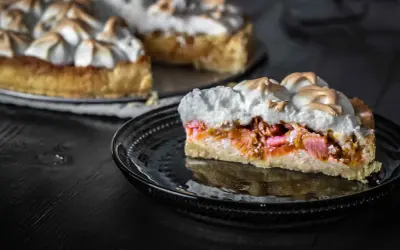 Ein Stück Rhabarberkuchen mit Baiser, das auf einem schwarzen Teller liegt. Der Kuchen hat eine goldbraune Kruste und eine saftige Rhabarberfüllung.