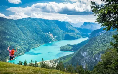 Giovane salta felice con zaino rosso, godendosi le passeggiate facili vicino a Trento con vista su lago turchese e montagne verdi.