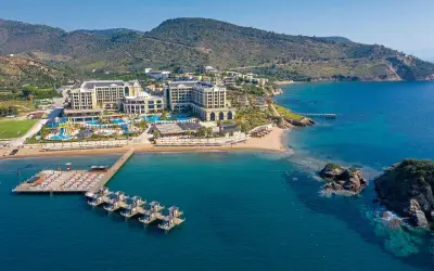 Luftaufnahme des Sune Efes Royal Palace mit Pools, Wasserrutschen und einem Steg mit Liegen am türkisfarbenen Meer.