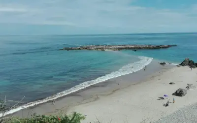 La **playa de Pechón** se extiende con arena dorada y aguas turquesas. Unas rocas emergen del mar, mientras algunas personas disfrutan del sol y el mar.